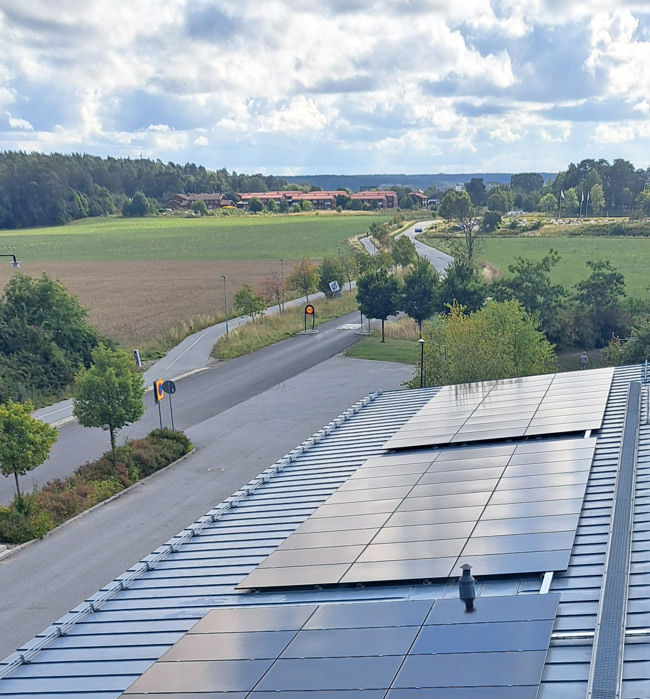 Solcellspaneler på taket av en skola med gröna åkrar och skog i bakgrunden.