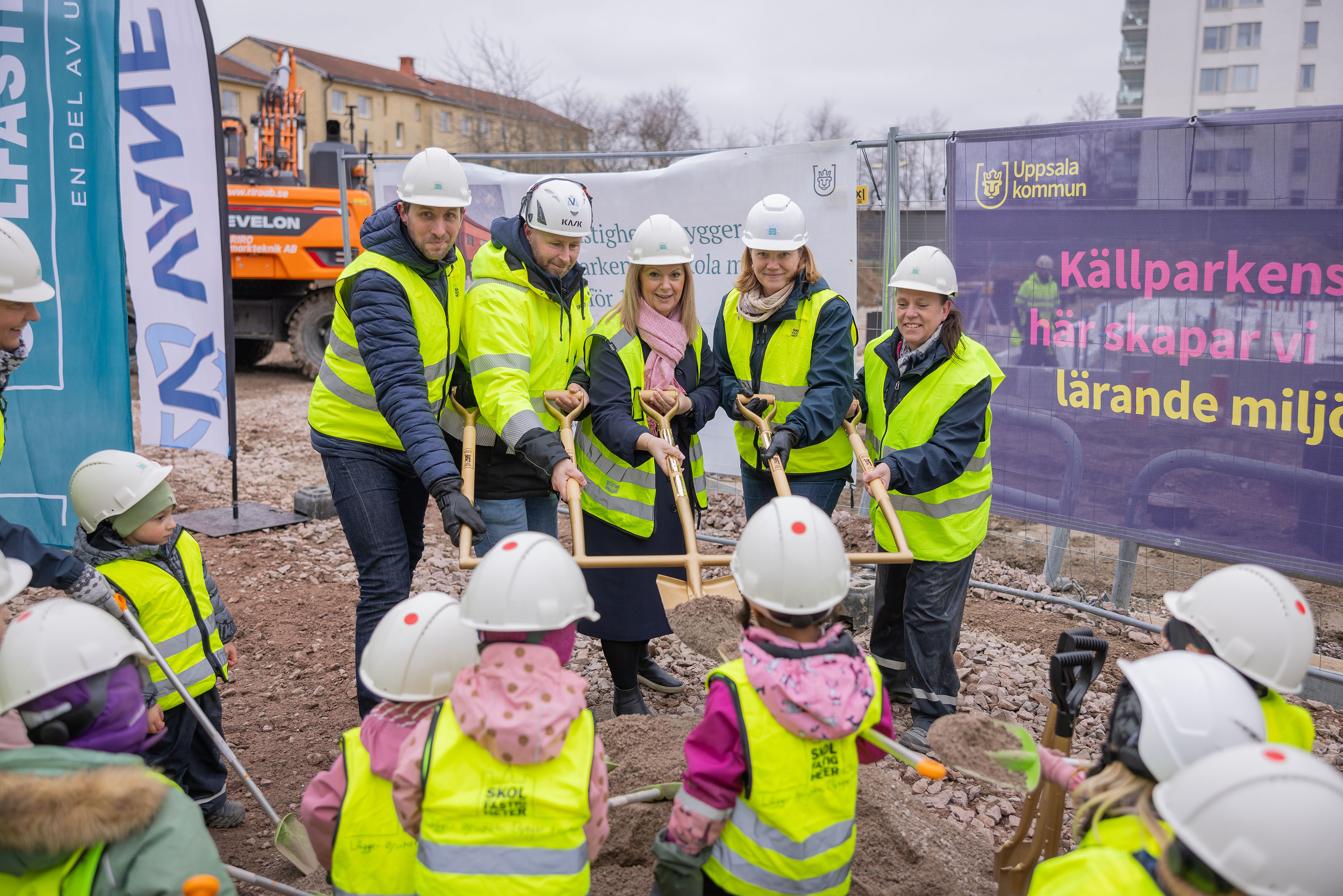 En grupp vuxna med hjälmar och västar håller i en gemensam spade på en byggplats. 
