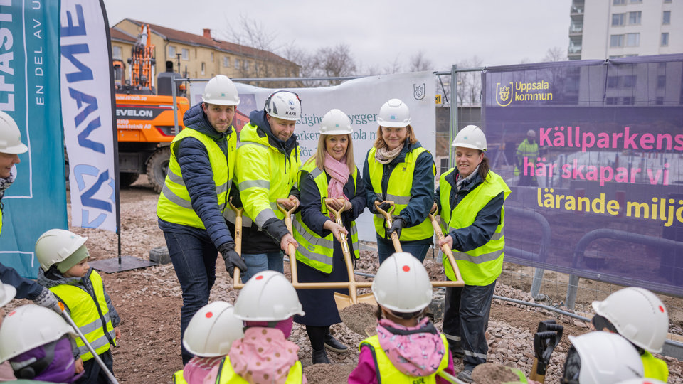 En grupp vuxna med hjälmar och västar håller i en gemensam spade på en byggplats. 