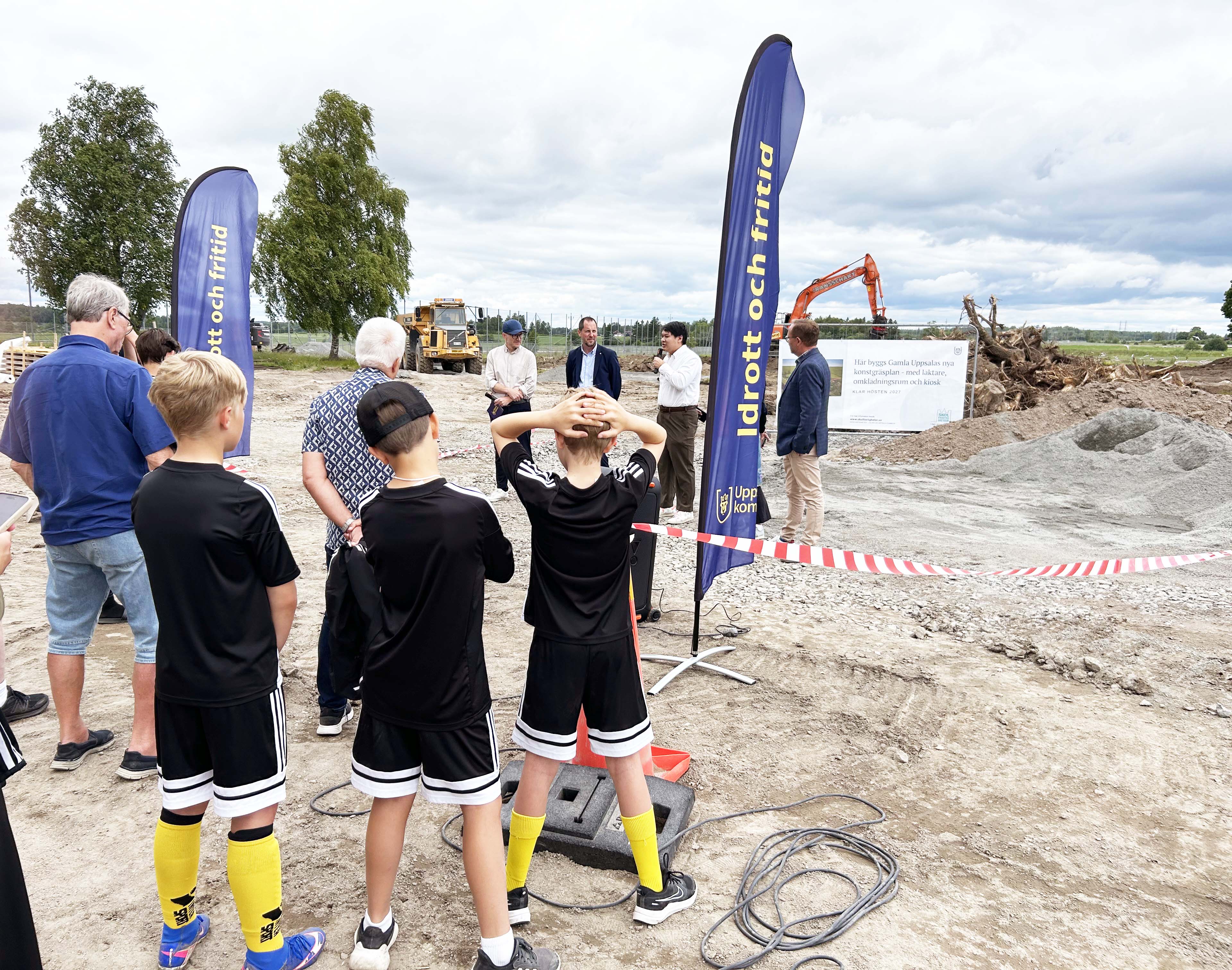 Ungdomar i fotbollskläder lyssnar på politiker som håller tal på en byggplats. 