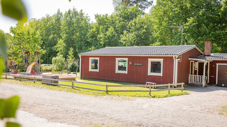 Röd förskola i trä med klätterställning och grön skog i bakgrunden.