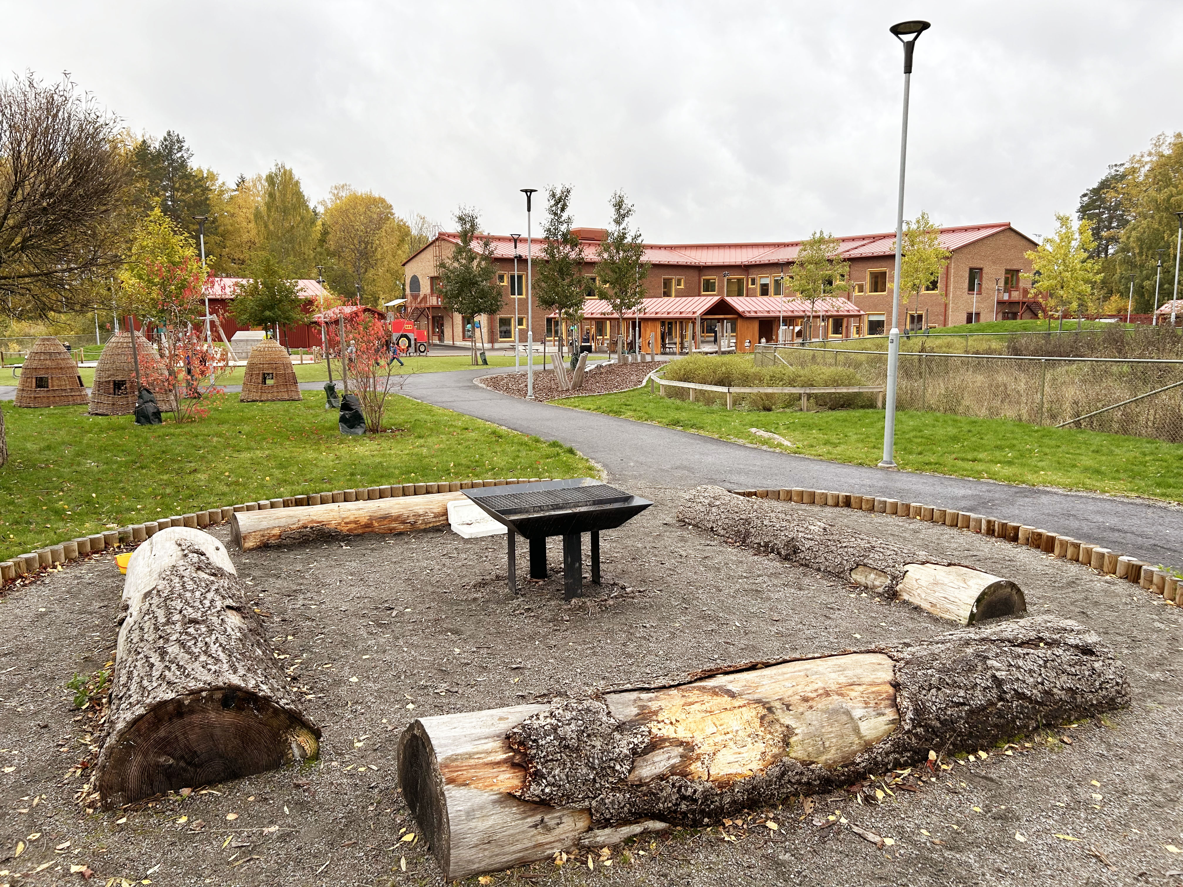Nybyggd förskola i rött tegel omgiven av höstskog och grillplats i förgrunden.