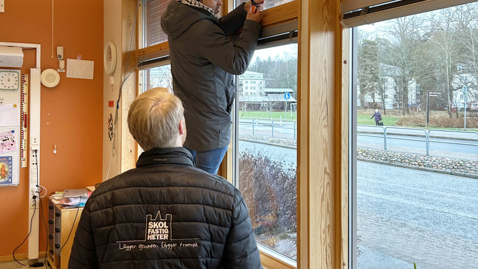 En person undersöker ett fönster i en skola framför en person som tittar på.