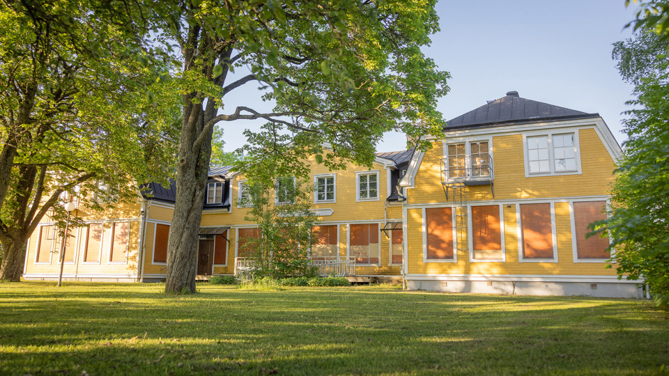 Gulmålad träbyggnad med grön gräsmatta och träd i förgrunden.