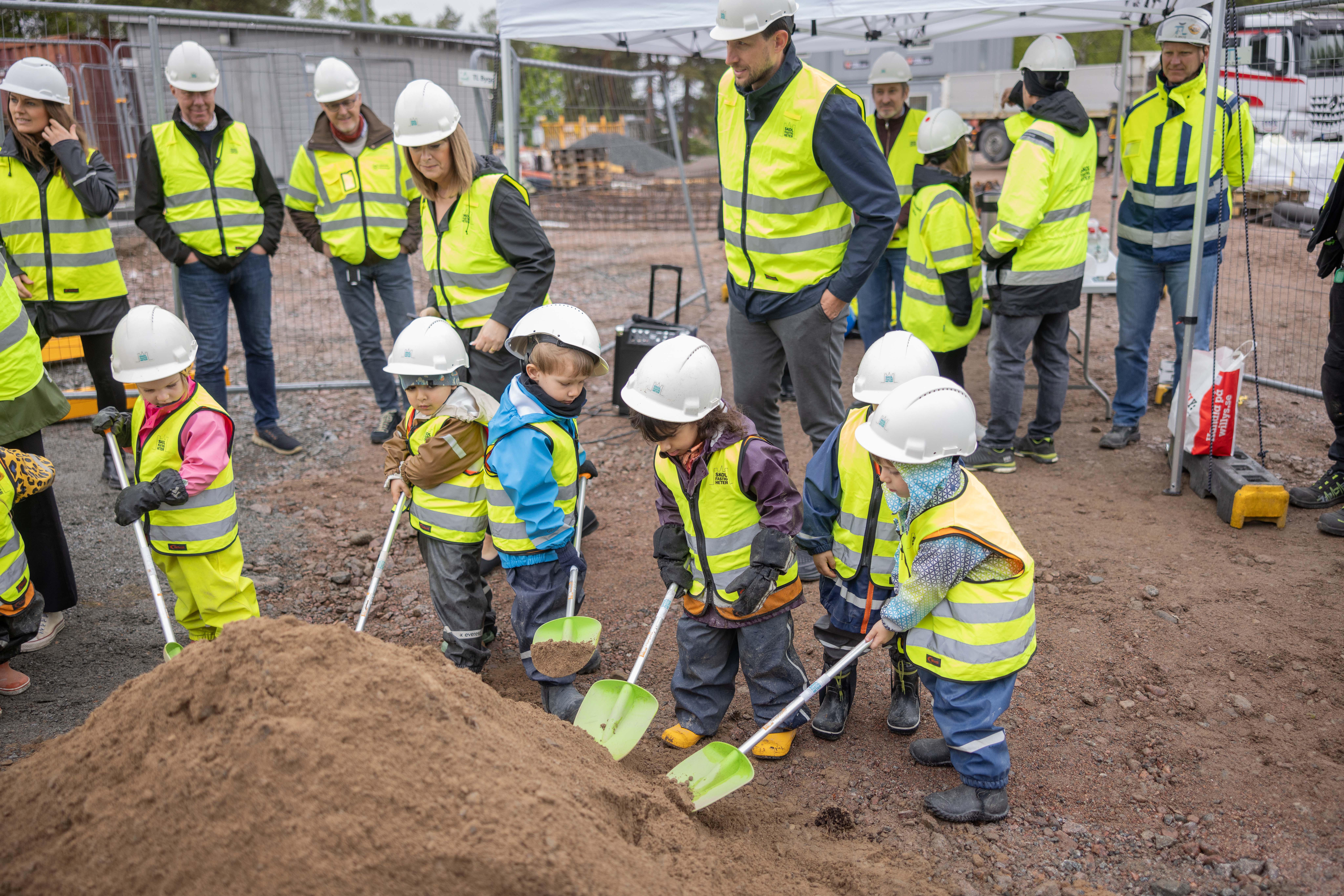 Förskolebarn i gula västar gräver i sandhög på en byggplats.