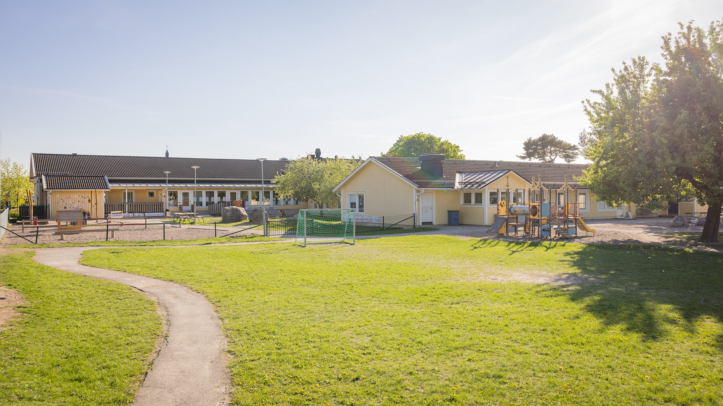 Gul förskola med förskolegård, klätterställning och gröna buskar i förgrunden.