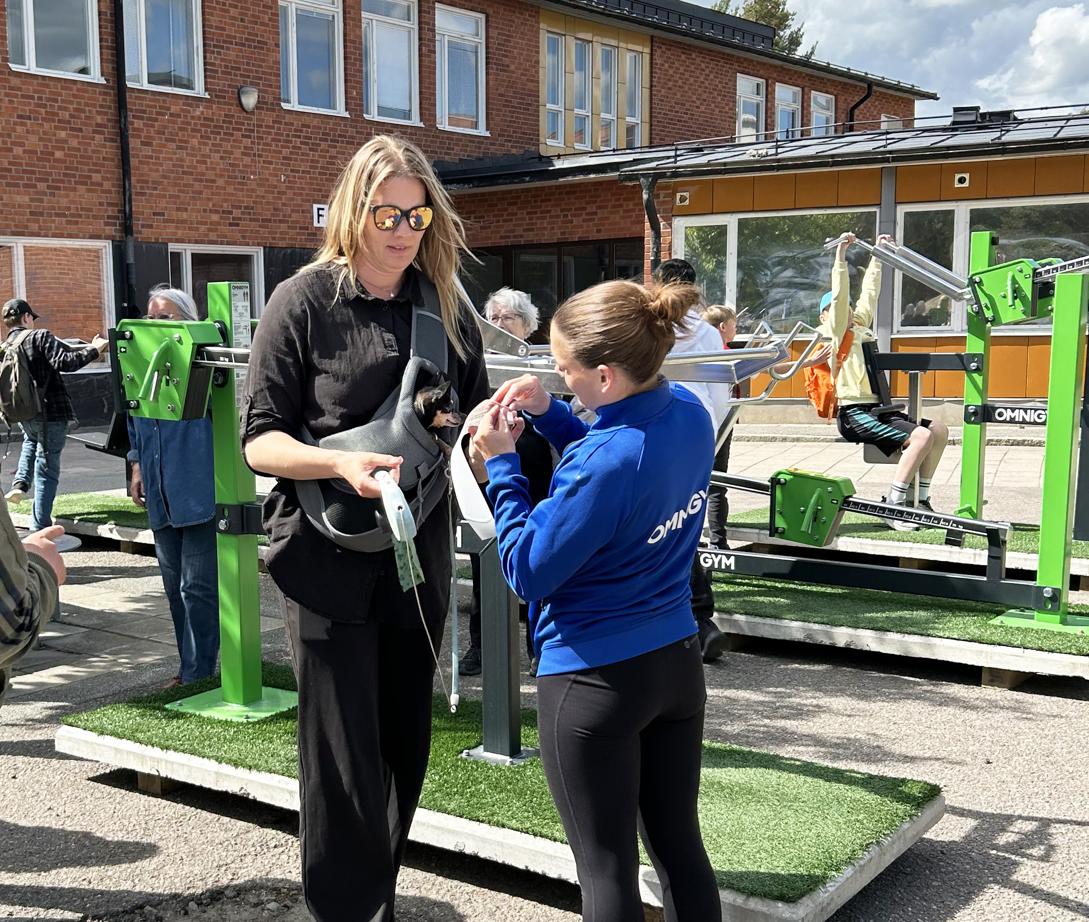 Person instruerar en kvinna vid ett utegym framför en skola.