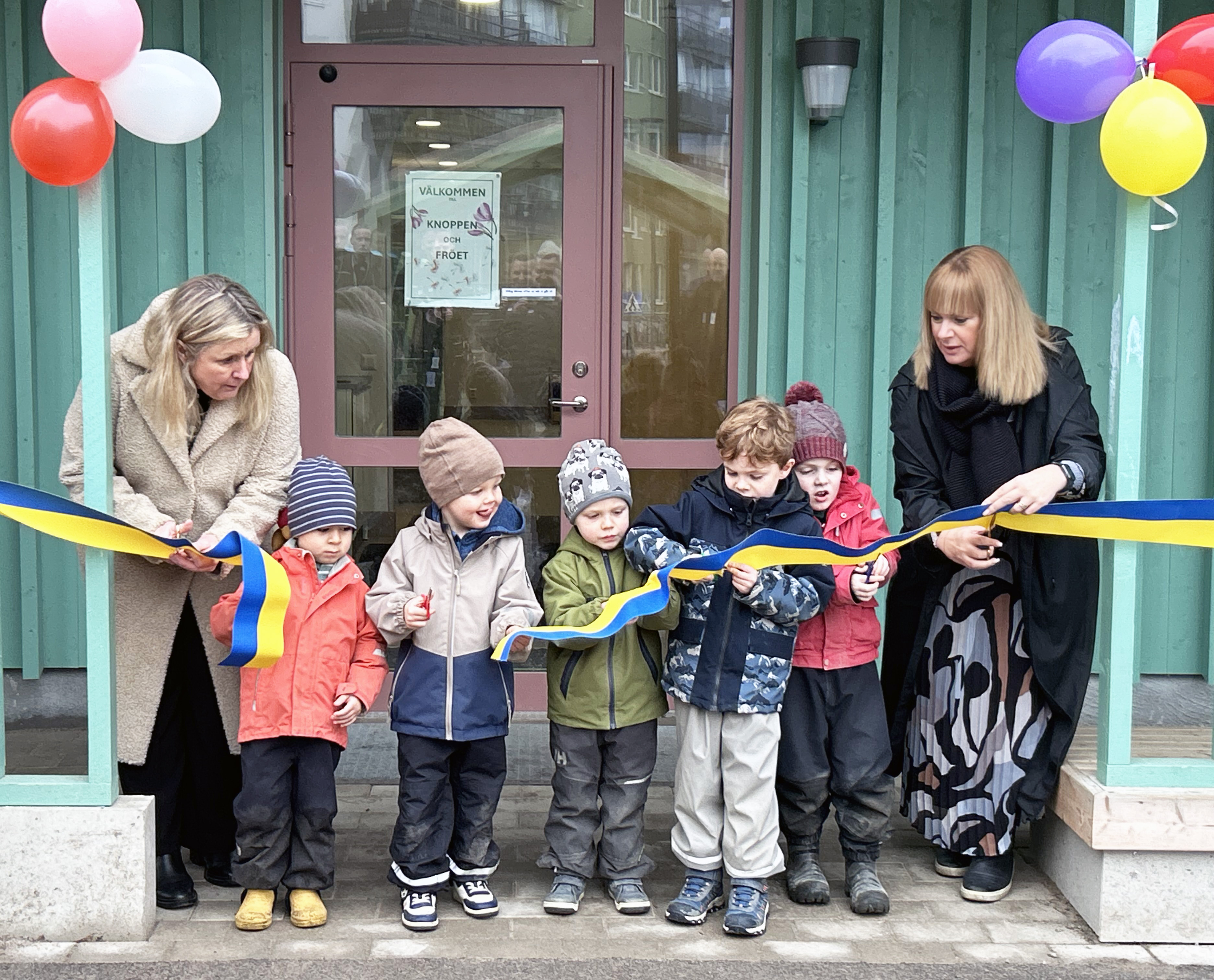 Förskolebarn och två vuxna klipper ett invigningsband framför en byggnad. Det finns ballonger runt dörren.