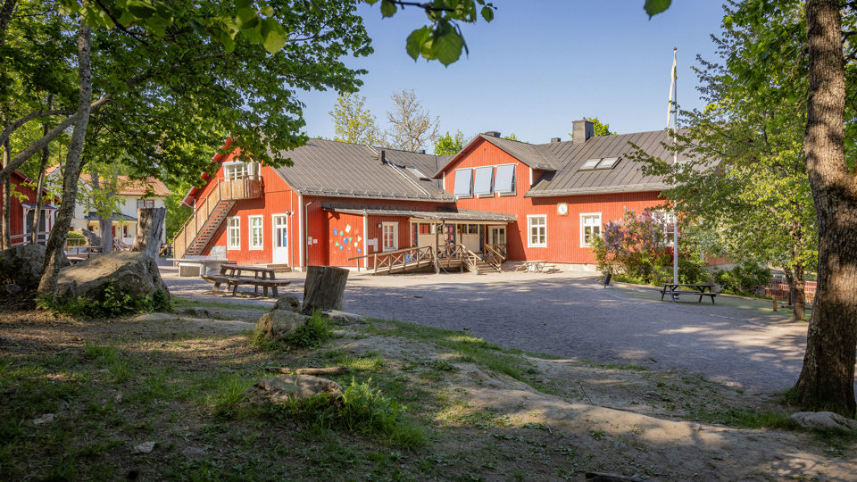 Röd skolbyggnad i trä, med grön skolgård och träbänkar i förgrunden