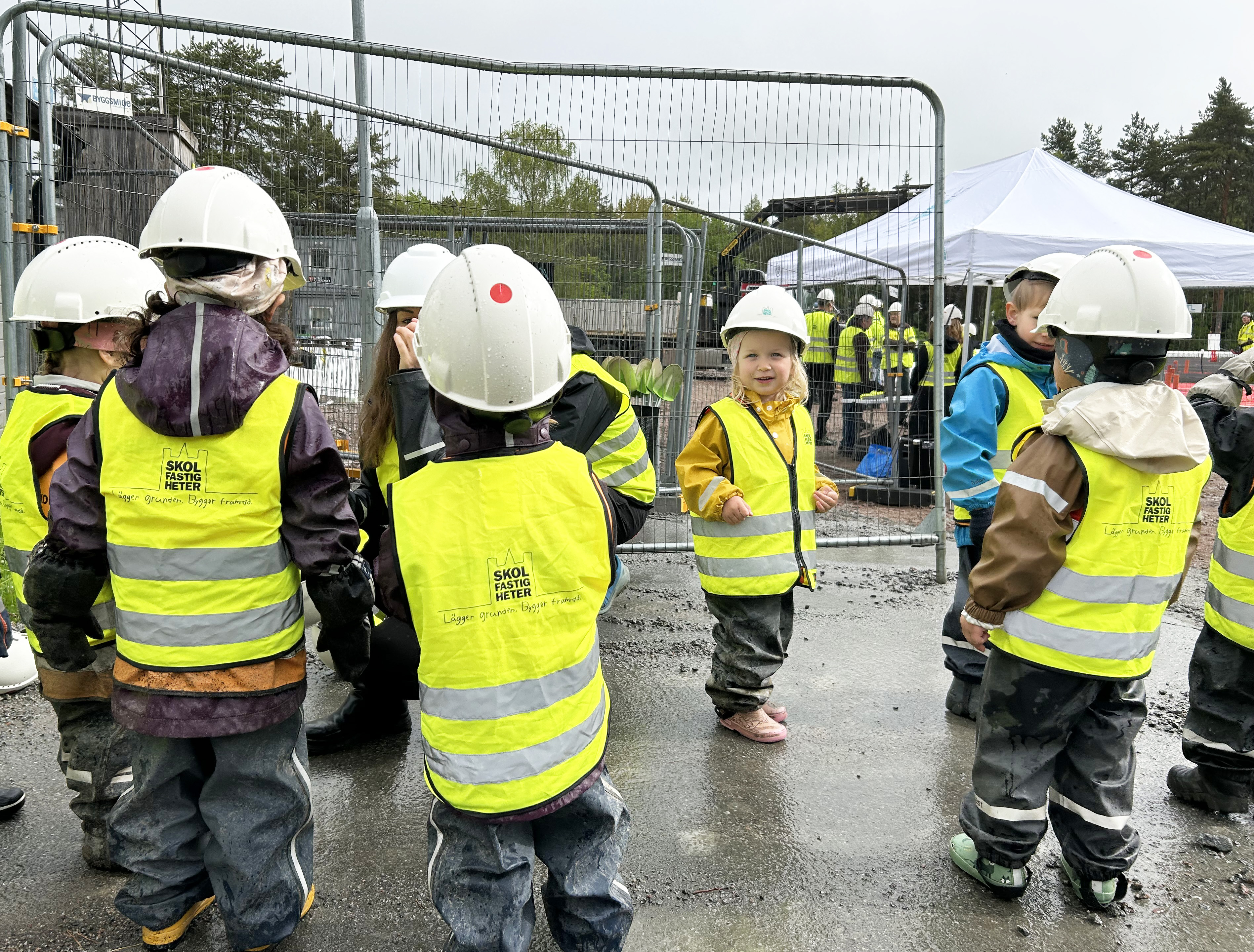 Förskolebarn i gula västar och bygghjälmar besöker en byggplats.