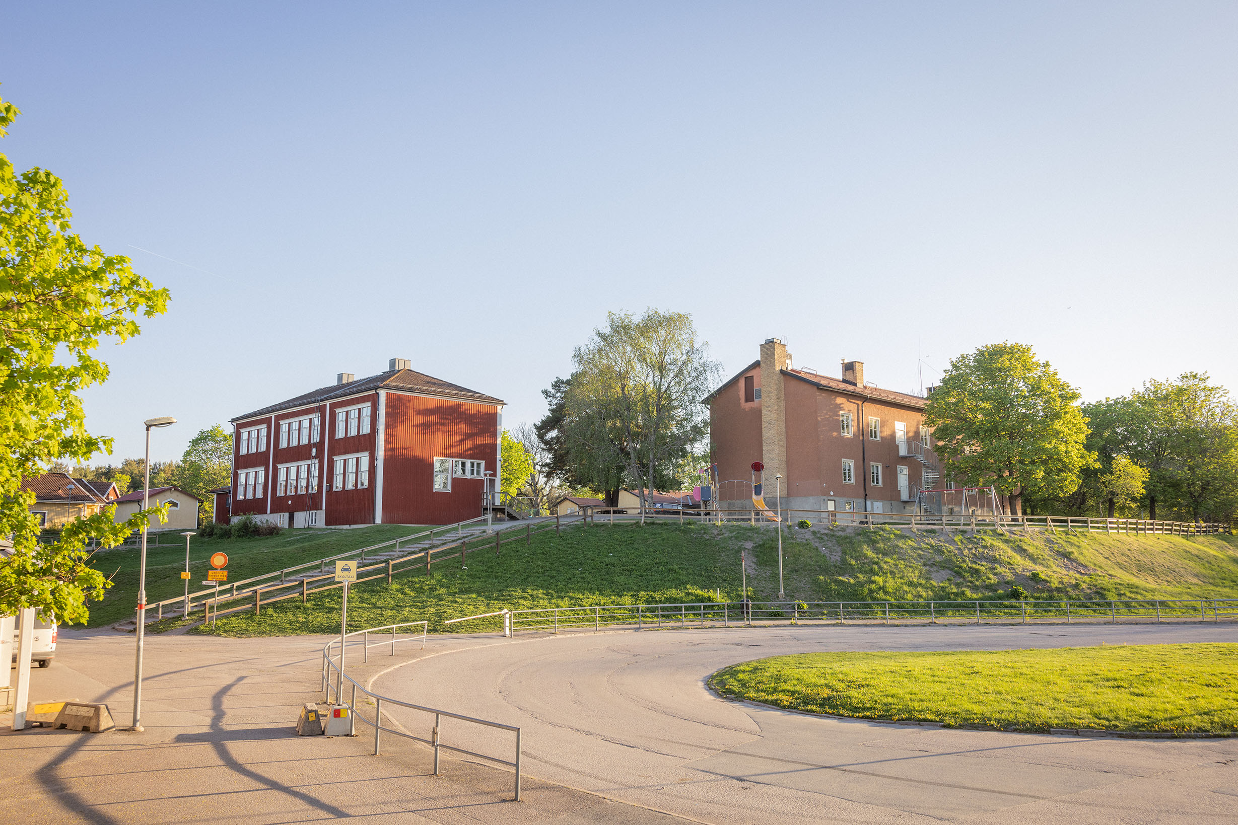 Röd skola i trä och brun byggnad i sten med en bilväg i förgrunden