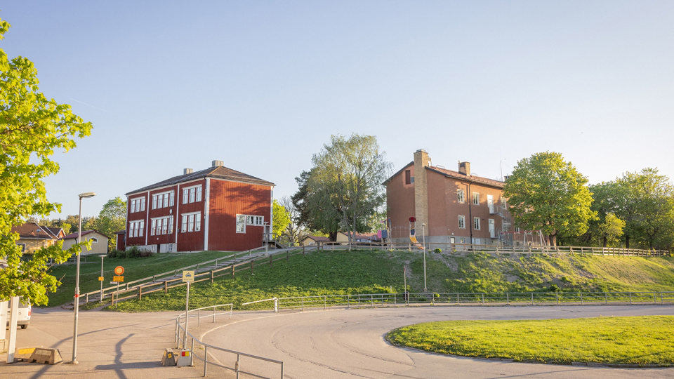 Röd skola i trä och brun byggnad i sten med en bilväg i förgrunden