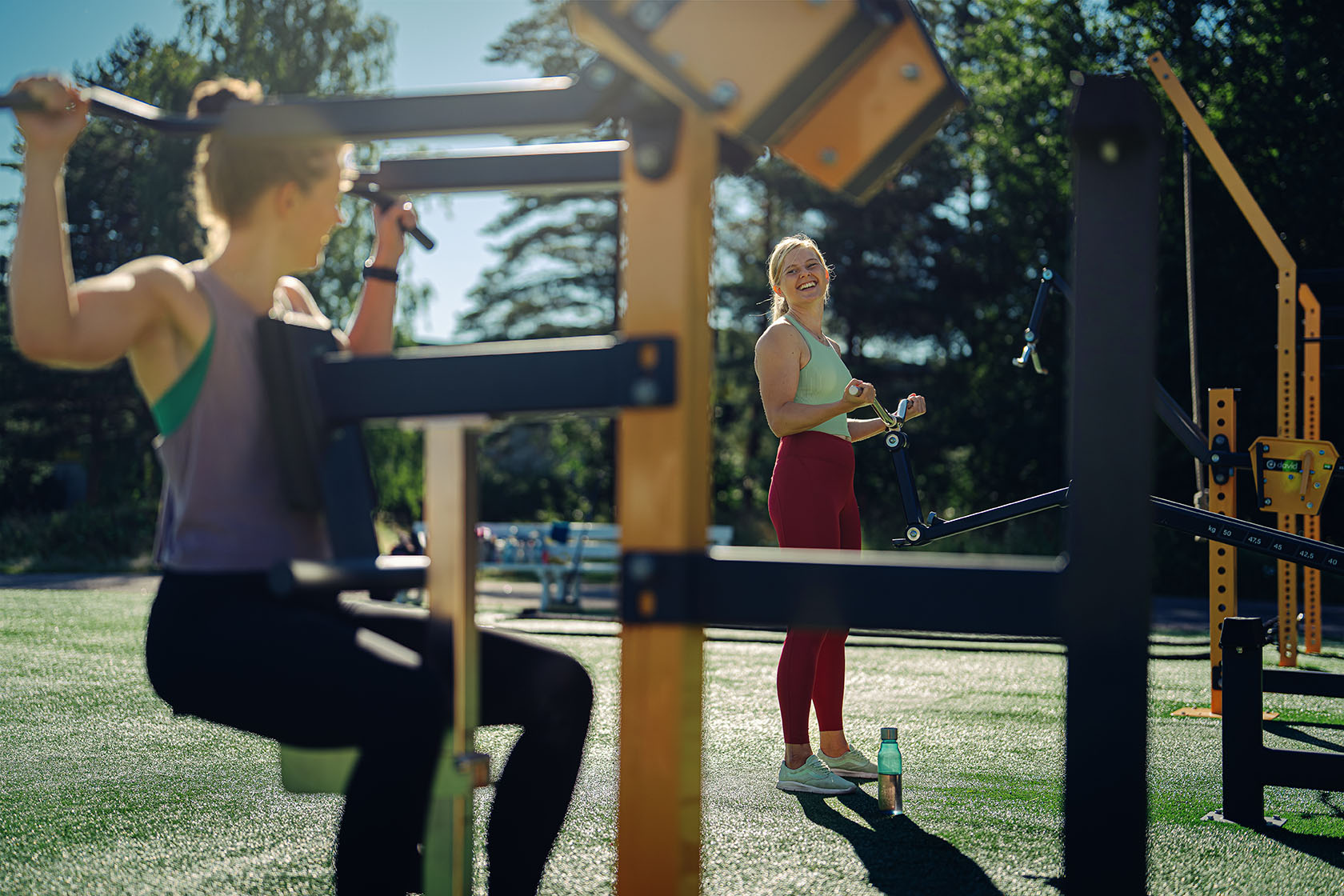Två unga kvinnor tränar på ett utegym i solen.