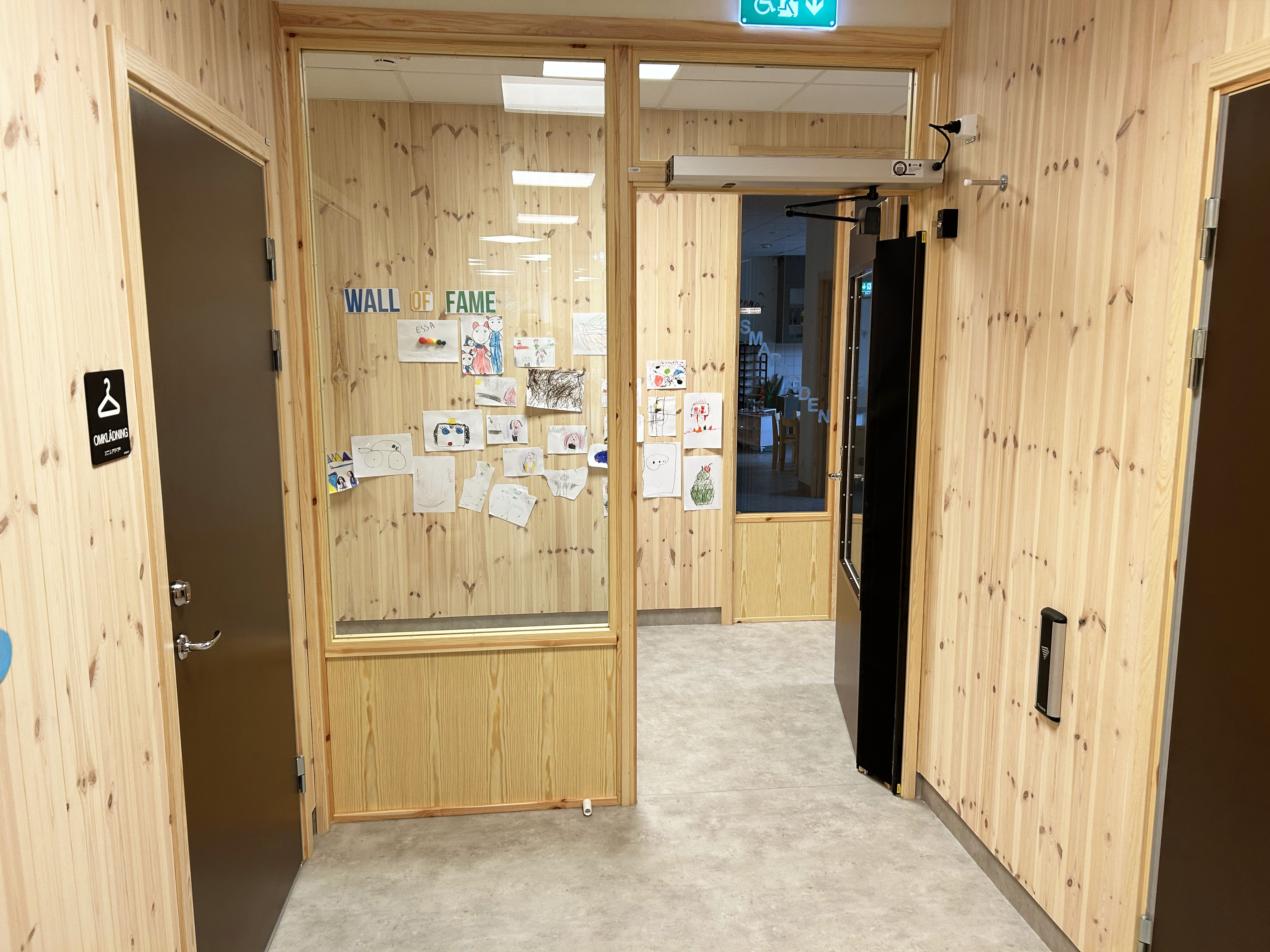 Träklädd korridor med öppna dörrar, anslagstavla och tillgänglighetsskylt.