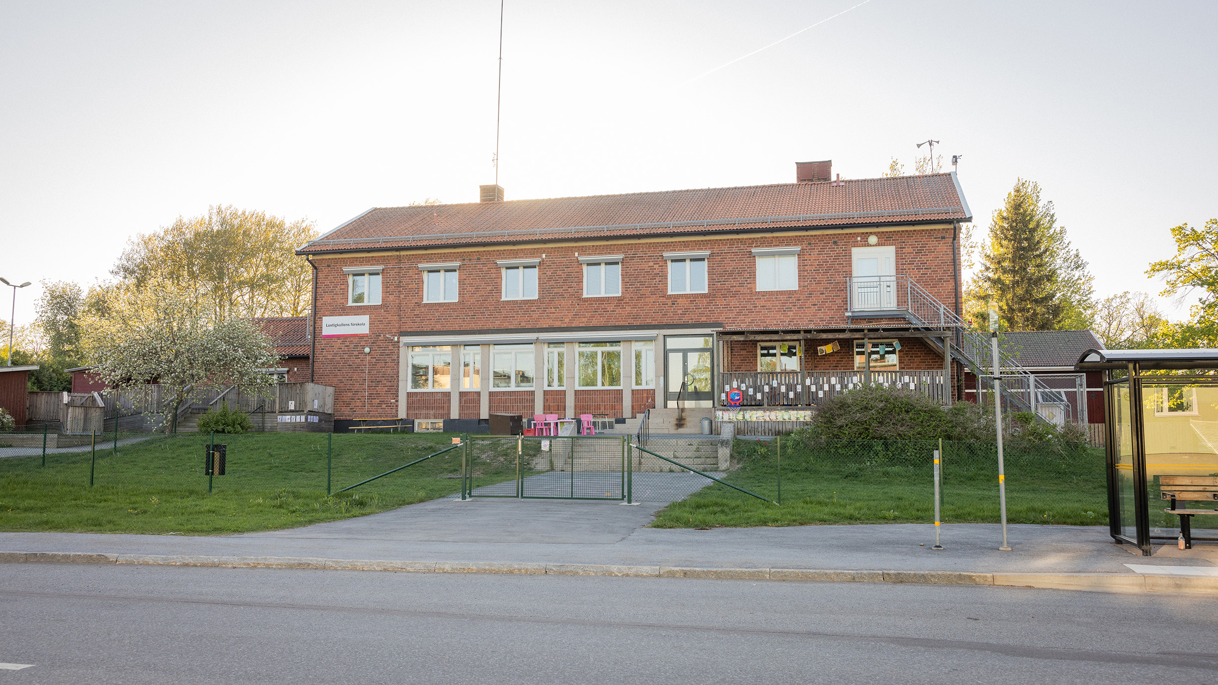 Brun tegelbyggnad med bilväg och busskur i förgrunden.