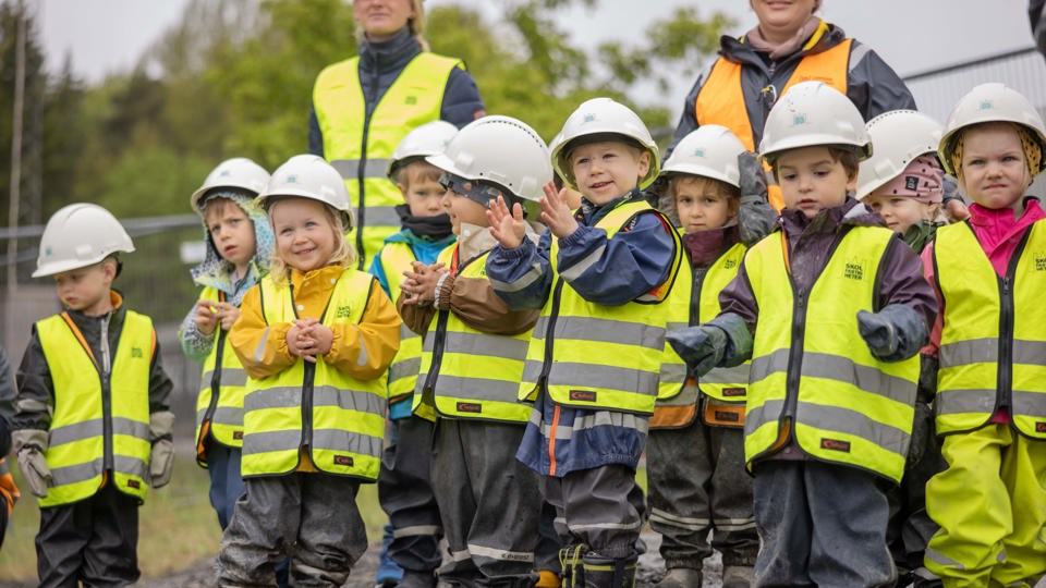 Förskolebarn i gula västar och bygghjälmar på besök vid en byggplats.